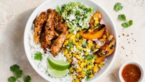 Chicken Taco Bowl with Grilled Peaches and Corn Salsa Chicken taco bowl topped with grilled peaches, corn salsa, mashed avocado, and cotija cheese in a white bowl