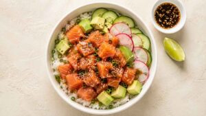 Overhead wide shot of a salmon avocado poke bowl with sushi rice, cubed avocado, cucumber, radish, and furikake