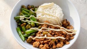 Overhead wide shot of a sesame chicken rice bowl with steamed rice, glazed chicken mince, green beans, and edamame