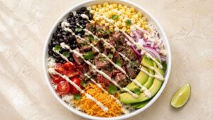 Steak taco bowl with sliced grilled steak, rice, avocado, black beans, corn, and lime crema